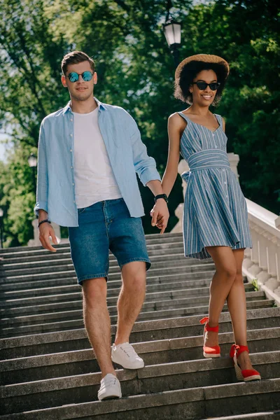 Young stylish couple in sunglasses holding hands and going downstairs in park — Stock Photo