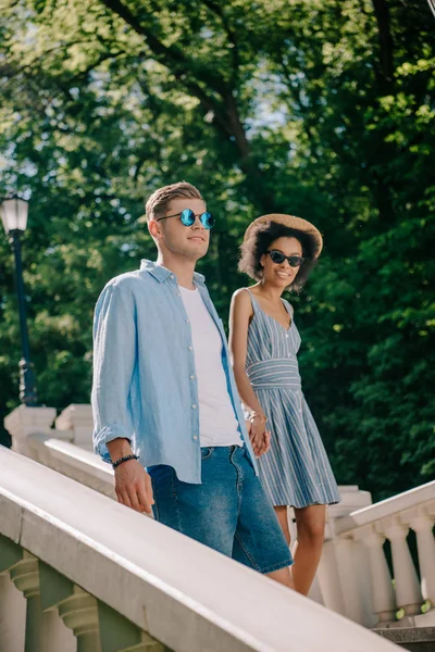 Sourire interracial couple en lunettes de soleil descendre dans le parc — Photo de stock