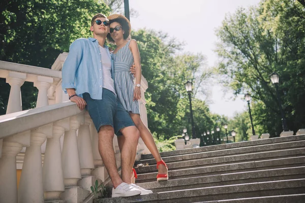 Vista basso angolo di elegante coppia multiculturale in piedi sulle scale nel parco — Foto stock