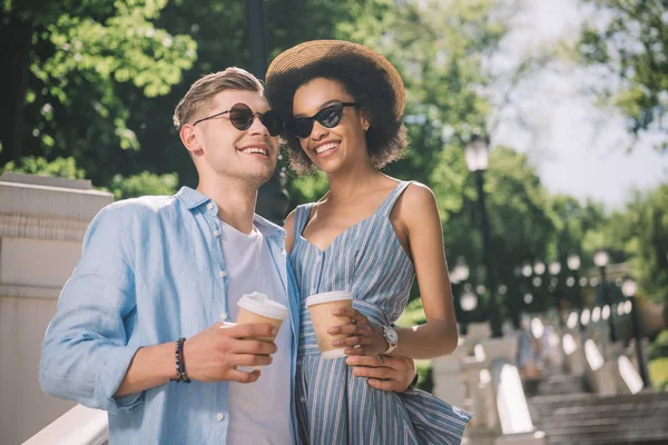 Feliz pareja multiétnica en gafas de sol con tazas de café de pie en las escaleras en el parque - foto de stock