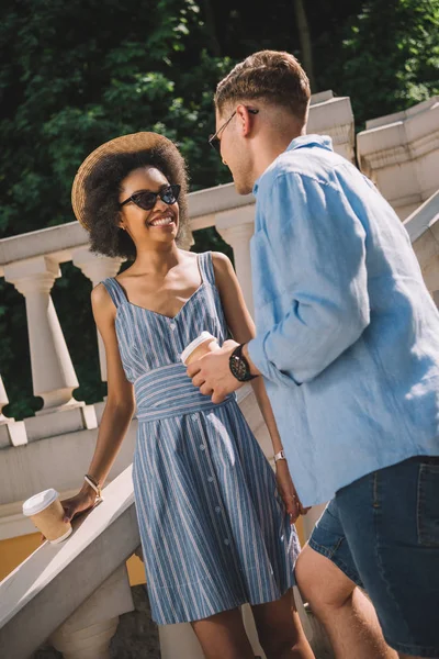 Casal interracial com copos de café descartáveis conversando uns com os outros — Fotografia de Stock