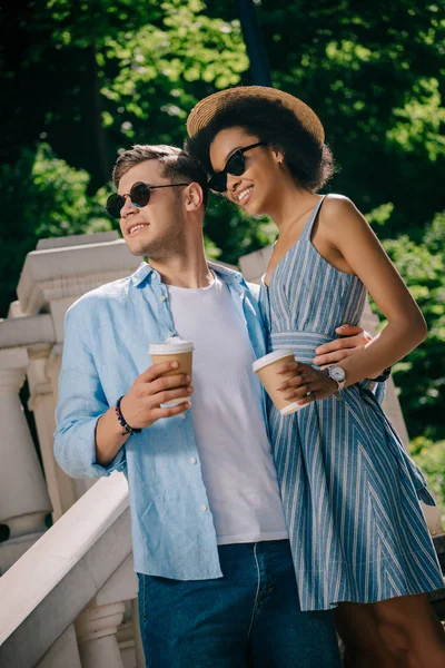 Interracial elegante pareja en gafas de sol con papeles tazas de café en las escaleras - foto de stock