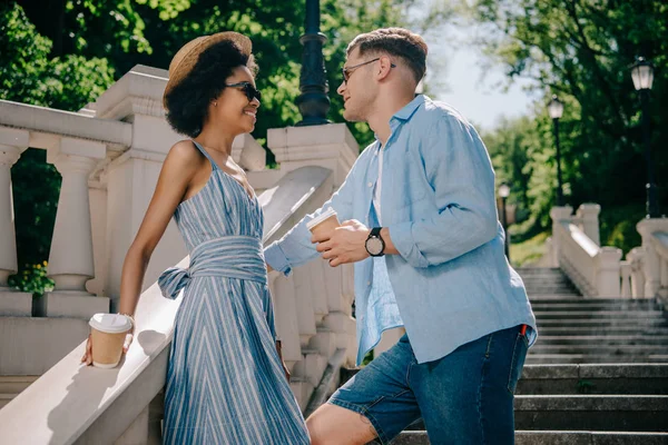 Vue latérale du couple multiculturel élégant avec des tasses à café debout sur les escaliers — Photo de stock