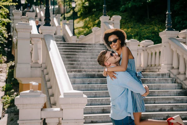 Glücklicher junger Mann hält afrikanisch-amerikanische Freundin in der Nähe von Treppen im Park — Stockfoto