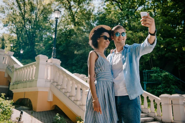 Low angle view of smiling multiethnic couple in sunglasses taking selfie on smartphone in park — Stock Photo