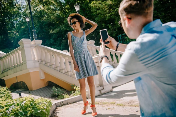 Hombre tomando la foto de la mujer afroamericana con estilo en el teléfono inteligente en el parque - foto de stock