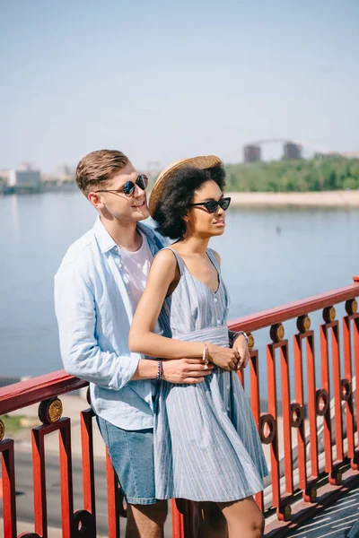 Pareja multicultural con estilo en gafas de sol de pie en el puente sobre el río - foto de stock