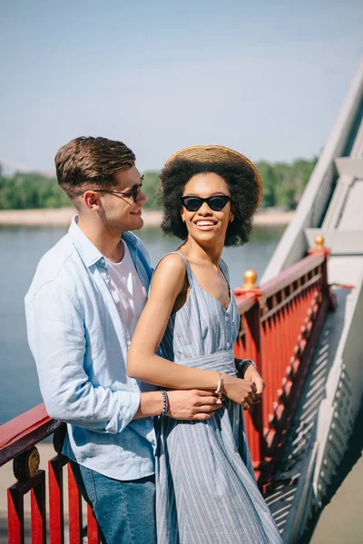 Joven elegante hombre en gafas de sol abrazando afrifcan american novia en puente - foto de stock