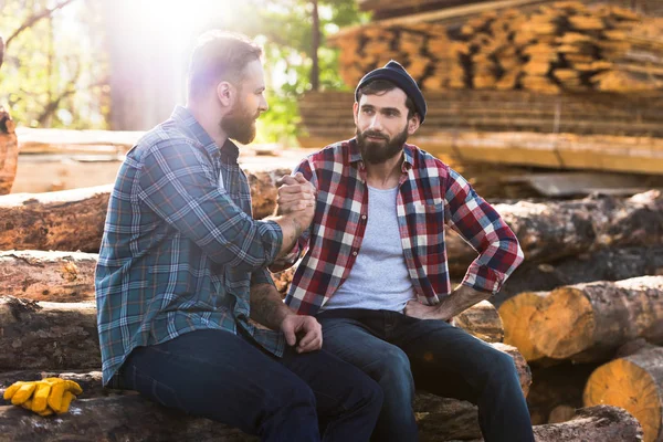 Bearded lumberjacks sitting on logs and shaking hands at sawmill — Stock Photo