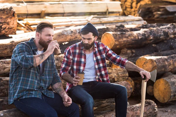 Two bearded loggers sitting with disposable cups of coffee and axe — Stock Photo