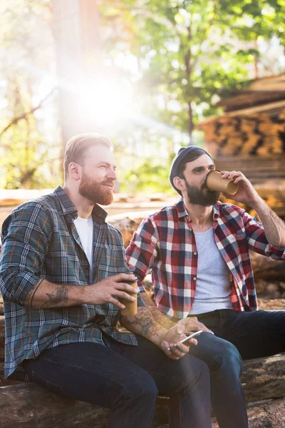 Bearded lumberjacks with smartphone drinking coffee from papers cups at sawmill — Stock Photo