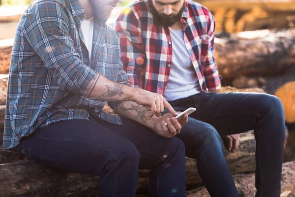 Cropped shot of bearded lumberjack pointing by finger on smartphone screen to partner — Stock Photo