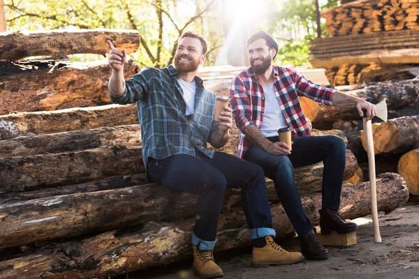 Lächelnder Holzfäller mit Kaffeetasse zeigt mit dem Finger auf Partner mit Axt, der auf Baumstämmen am Sägewerk sitzt — Stockfoto