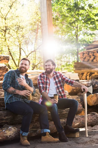 Holzfäller mit Kaffeetasse zeigt mit dem Finger auf Partner mit Axt, der auf Baumstämmen am Sägewerk sitzt — Stockfoto