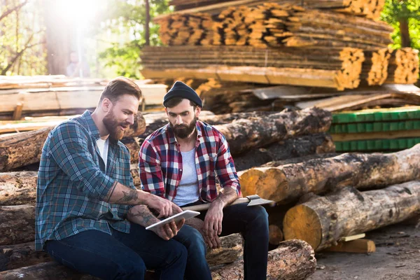 Bärtige Holzfäller sitzen mit Axt und digitalem Tablet auf Baumstämmen im Sägewerk — Stockfoto