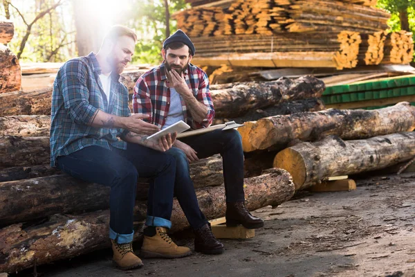 Two lumberjacks sitting on logs with axe and using digital tablet at sawmill — Stock Photo