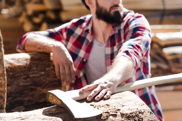 Cropped image of bearded lumberjack in checkered shirt with axe at sawmill — Stock Photo