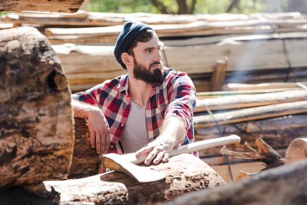 Bearded lumberjack in checkered shirt with axe looking away at sawmill — Stock Photo