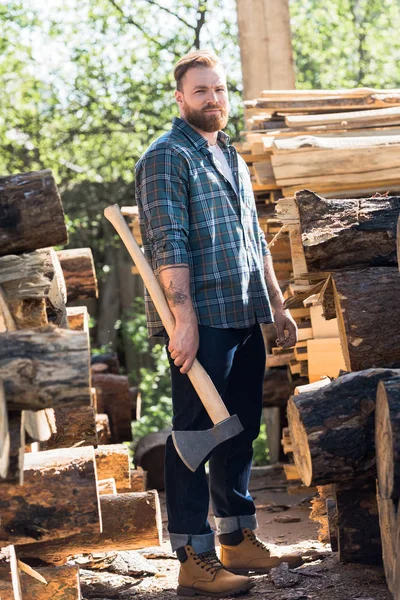 Bearded lumberjack  in checkered shirt standing with axe at sawmill — Stock Photo