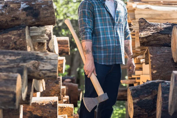 Cropped image of lumberjack in checkered shirt with tattooed hands holding axe between logs at sawmill — Stock Photo