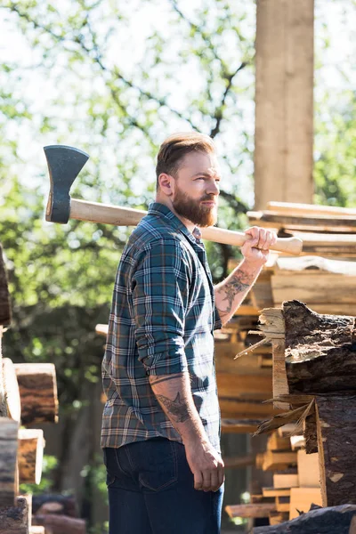 Bearded lumberjack in checkered shirt with tattooed hand holding axe on shoulder at sawmill — Stock Photo