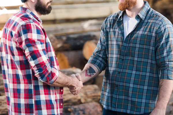 Tiro recortado de dos leñadores barbudos en camisas a cuadros estrechando la mano en el aserradero - foto de stock