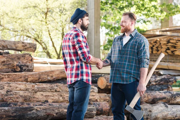 Side view of smiling lumberjack with tattooed hand holding axe and shaking hands with partner at sawmill — Stock Photo