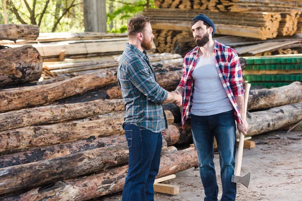 Lumberjack with holding axe and shaking hands with partner at sawmill — Stock Photo