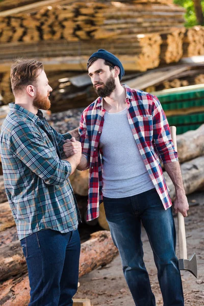 Bearded lumberjack in checkered shirt holding axe and shaking hands with partner at sawmill — Stock Photo