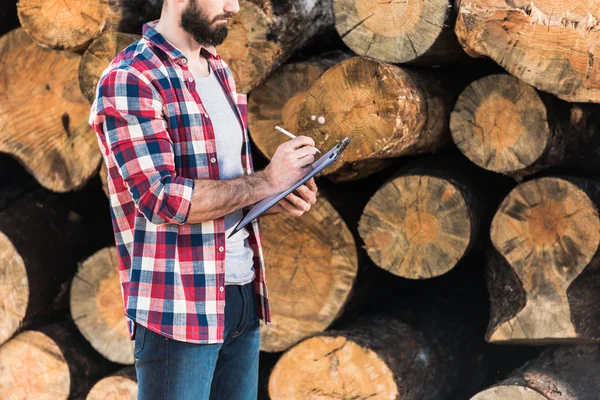 Schnappschuss eines bärtigen Holzfällers in kariertem Hemd, Schrift in Klemmbrett auf Baumstämme Hintergrund — Stockfoto