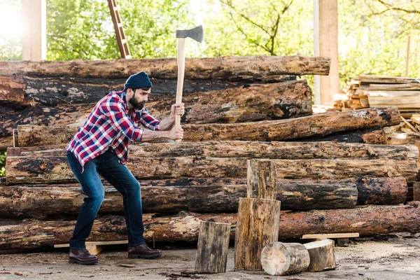 Side view of bearded lumberjack in checkered shirt chopping log at sawmill — Stock Photo