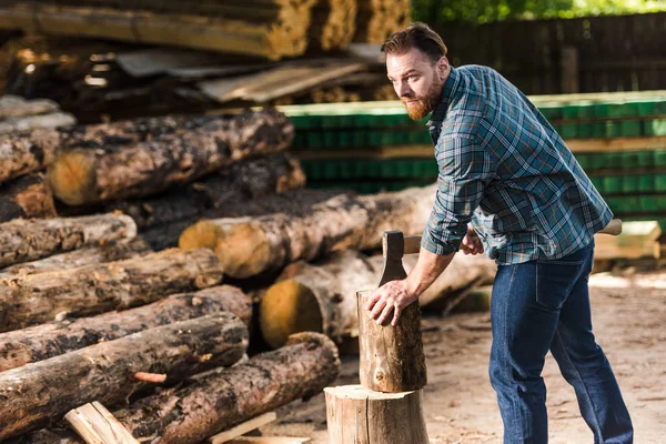 Bearded lumberjack in checkered shirt chopping log at sawmill — Stock Photo