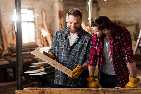 Happy carpenter in protective gloves showing wooden plank to partner at sawmill — Stock Photo