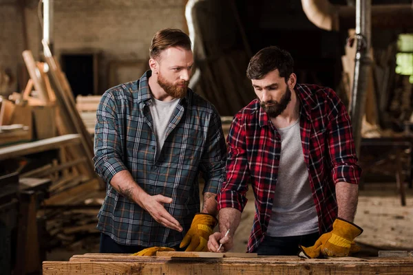 Carpenter in protective gloves pointing on wooden plank to partner with pen in hand at sawmill — Stock Photo