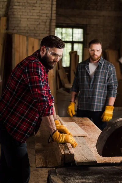 Bearded carpenter in safety googles using machine saw and partner standing behind at sawmill — Stock Photo