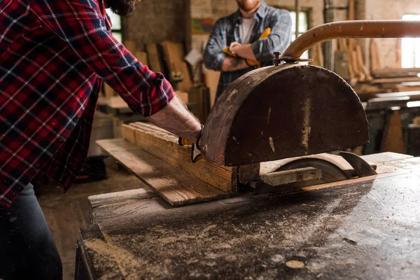 Cropped shot of carpenter in checkered shirt using machine saw and partner standing behind at sawmill — Stock Photo