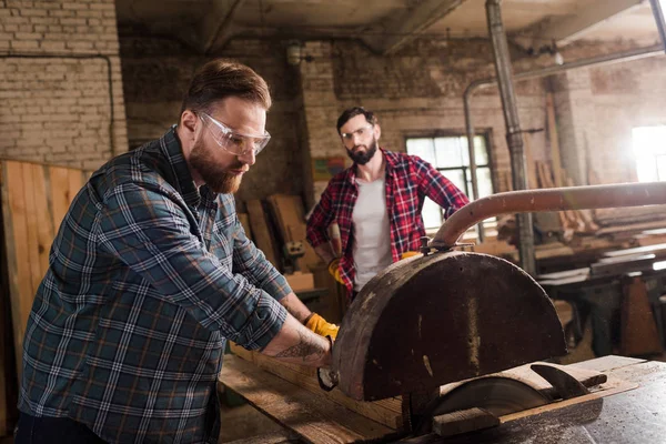Bearded carpenter in safety googles using machine saw and partner with crossed arms standing behind at sawmill — Stock Photo