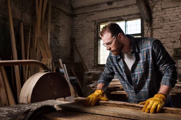 Carpinteiro em luvas de proteção e óculos usando serra de máquina na serraria — Fotografia de Stock