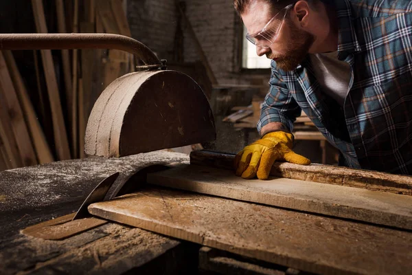 Tiro cortado de carpinteiro barbudo em luvas de proteção e óculos usando serra de máquina na serraria — Fotografia de Stock