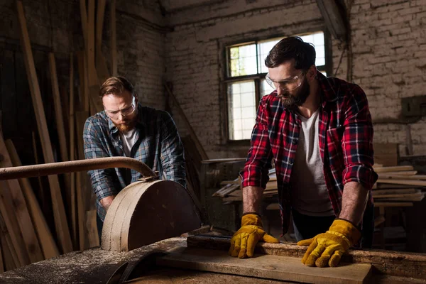 Bearded carpenter in protective googles and gloves using machine saw and partner standing near at sawmill — Stock Photo