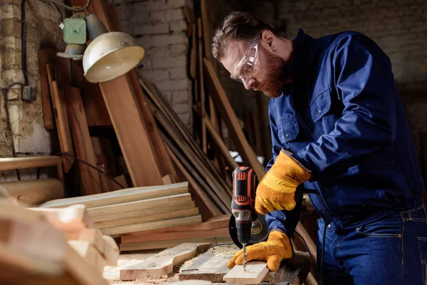 Bearded carpenter in protective gloves and googles using electric drill at sawmill — Stock Photo