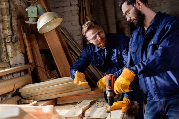 Bearded craftsman pointing to partner in protective gloves and googles working with electric drill at sawmill — Stock Photo