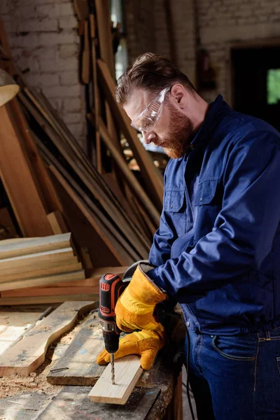 Side view of bearded worker in protective gloves and googles using electric drill at sawmill — Stock Photo