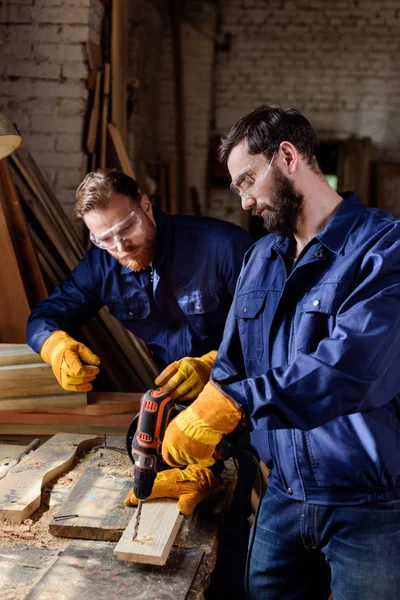 Artesano señalando a la pareja en guantes de protección y googles trabajando con taladro eléctrico en carpintería - foto de stock