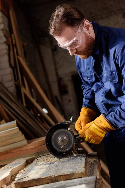 Bearded craftsman in protective googles and gloves using grinding machine at sawmill — Stock Photo