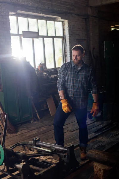 Bearded craftsman in protective gloves standing near machine at sawmill — Stock Photo