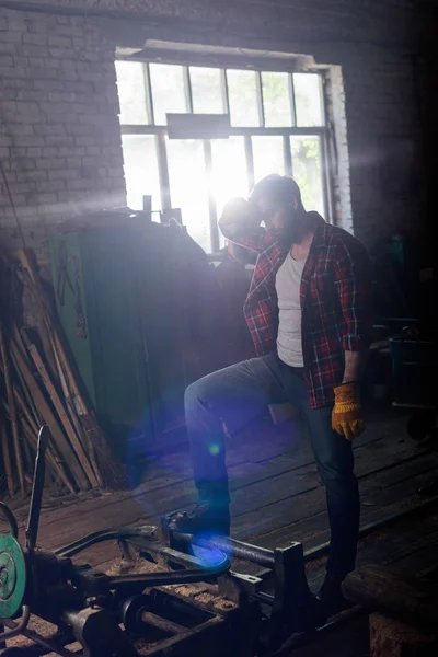 Bearded worker in protective gloves wiping forehead and standing near machine at sawmill — Stock Photo
