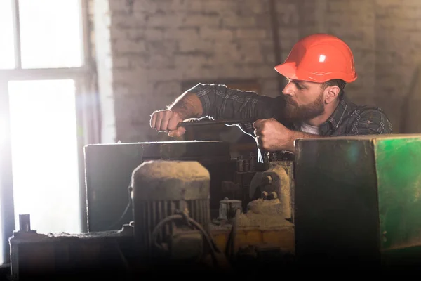 Bearded worker in protective helmet repairing machine tool at sawmill — Stock Photo