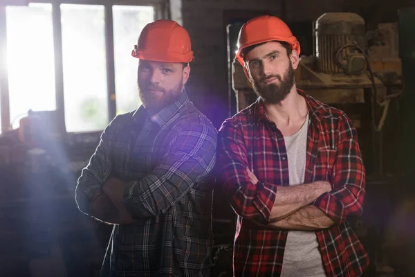 Two bearded carpenters with crossed hands in protective helmets standing at sawmill — Stock Photo