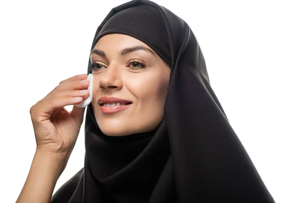 Sorridente jovem muçulmana no hijab esfregando rosto com almofada de algodão isolado no branco — Fotografia de Stock